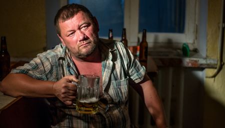 Мужчина в состоянии алкогольного опьянения сидит за столом и держит кружку пива